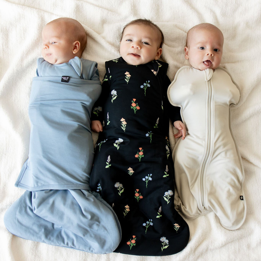 Three babies in sleep bags and swaddles on a cream blanket
